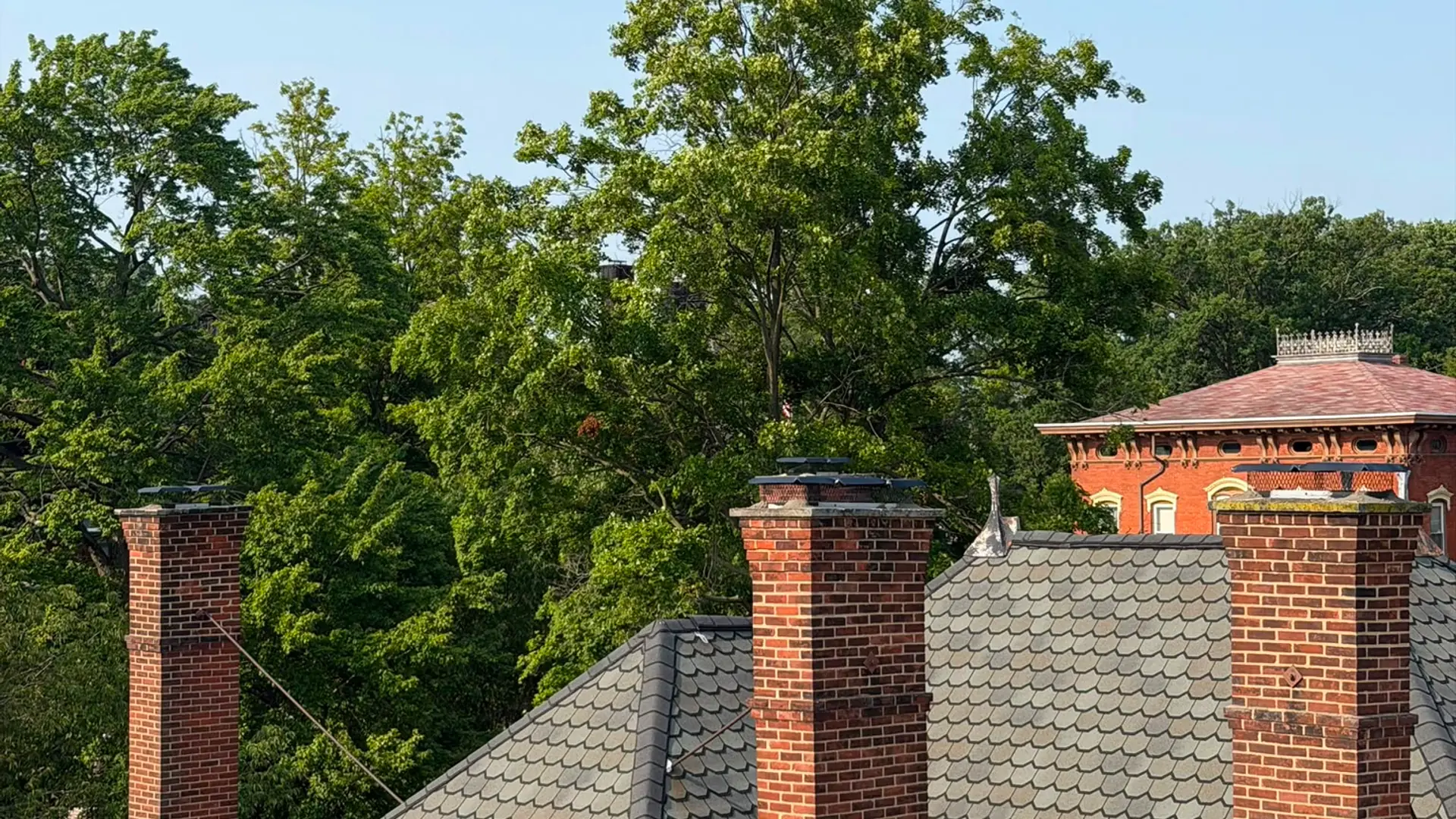 Three brick chimneys rising from the slate roof of an older brick home surrounded by mature trees